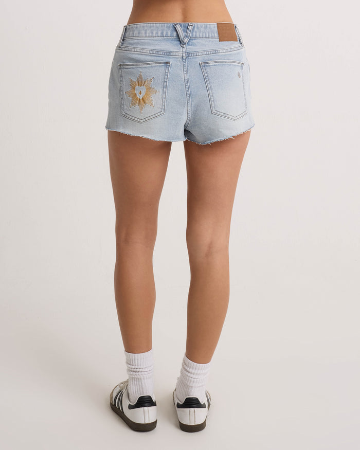 A person faces away, wearing Volcom Women's Cruz Control light blue high-rise denim shorts in chambray wash with frayed hems and a sun patch on the pocket, paired with white socks and black-and-white sneakers.