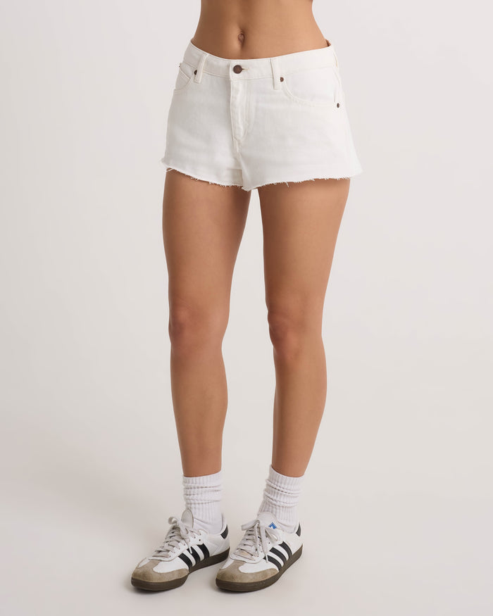 A person wearing Volcom Women's Cruz Control vintage white high-rise denim shorts, white socks, and black-and-white Adidas sneakers stands against a plain white background. Only the lower half of the body is visible.
