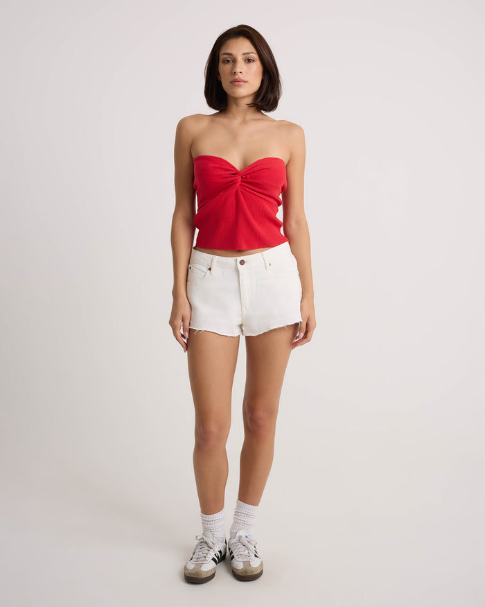 A woman faces forward wearing the Volcom Women's Cruz Control red strapless top and vintage white high-rise denim shorts, styled with white socks and sneakers, against a plain light background.
