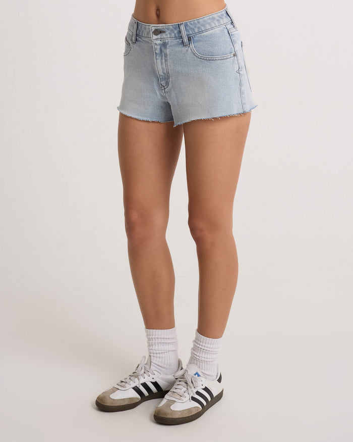 A person wears Volcom Women's Cruz Control light blue chambray wash denim shorts, white socks, and white Adidas sneakers with black stripes, standing against a plain white background. Only the lower half of their body is visible.