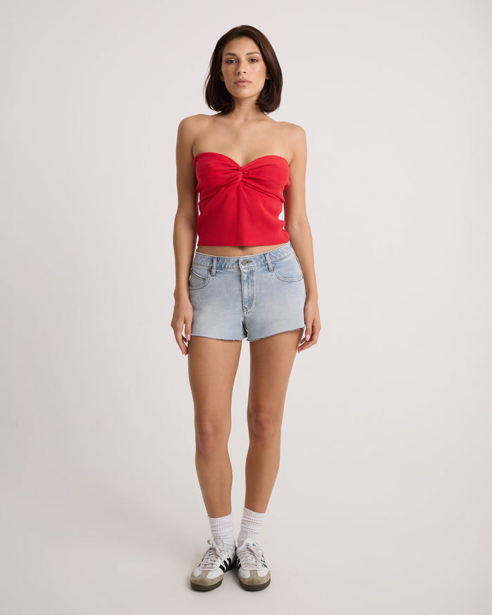 A person stands facing forward in the Volcom Women's Cruz Control red strapless top and light blue high-rise chambray-wash denim shorts, with white socks and black-and-white sneakers, against a plain white background.