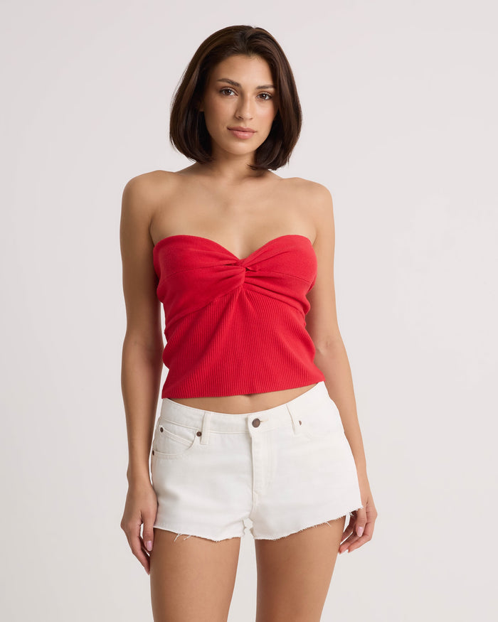 A woman wearing the Volcom Women's Cruz Control red strapless top with twisted knot detail and vintage white high-rise denim shorts stands against a white background.