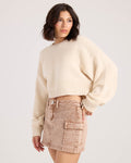 A woman with shoulder-length dark hair wears the Volcom Women's Dayz Like This Sweater and a light brown denim mini skirt with front pockets, standing against a plain light background.