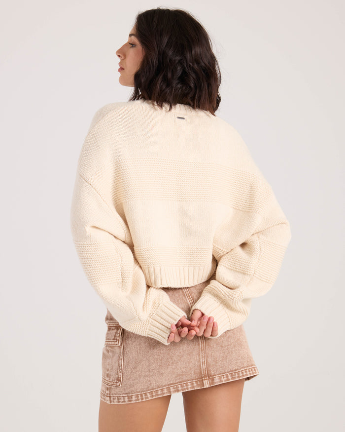 A woman with shoulder-length dark hair models the Volcom Women's Dayz Like This Sweater in white, standing with her back to the camera and hands clasped behind her.