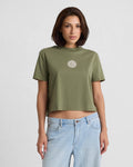 A woman wears the Volcom Women's Dial Crop Tee, a loose-fitting olive green organic cotton top with a white sun graphic, paired with light blue jeans, standing against a plain white background.