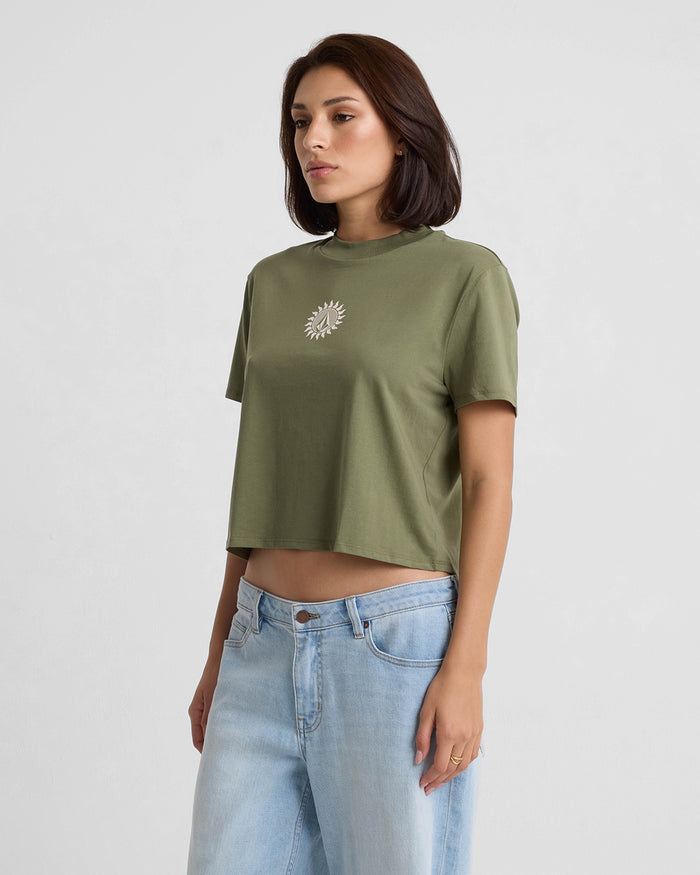 A woman with short dark hair and a neutral expression wears the Volcom Women's Dial Crop Tee, featuring short sleeves, organic cotton, olive green color, and white sun design, paired with light blue jeans against a plain light gray background.