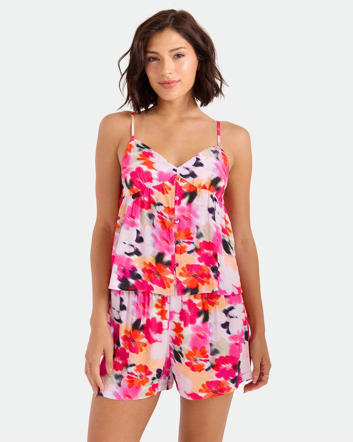 A woman models the Volcom Women's Dream In Bloom Cami, a sleeveless, floral-print top in an orange-blue palette with adjustable straps, standing against a plain light background and gazing at the camera with a relaxed look.