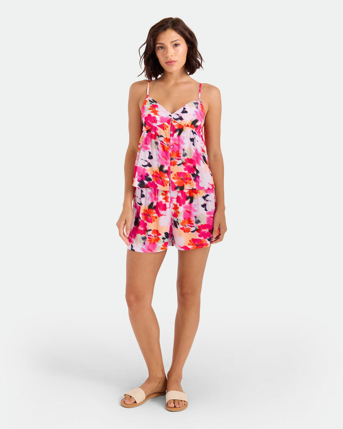 A woman with short dark hair and a relaxed expression stands against a white background wearing the Volcom Women's Dream In Bloom Cami floral romper in orange-blue tones, paired with tan flat sandals.