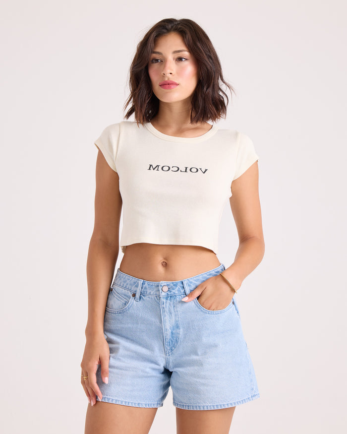 A woman with short brown hair wears the Volcom Women's Flip Side 1991 Mid Tee in vintage white, featuring a backward VOLCOM graphic, paired with light blue high-waisted denim shorts. She stands with one hand in her pocket against a plain light background.