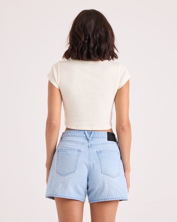 A woman with short dark hair is standing with her back to the camera, wearing the Volcom Women's Flip Side 1991 Mid Tee in vintage white and light blue denim shorts against a plain light background.