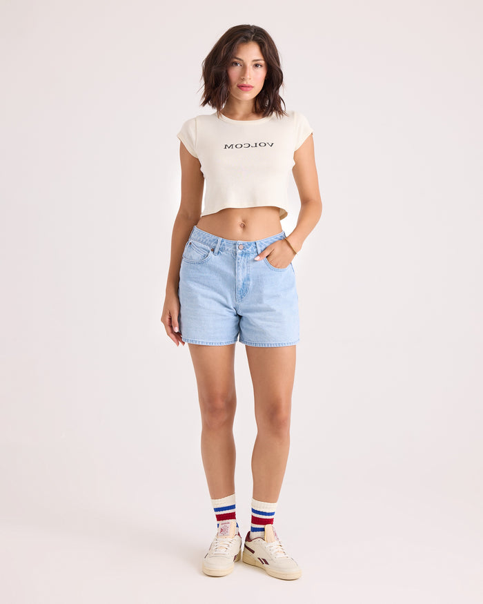A woman stands against a plain background wearing the Volcom Women's Flip Side 1991 Mid Tee—a vintage-inspired, fitted white cropped shirt—paired with light blue denim shorts, white sneakers, and white socks featuring red and blue stripes.