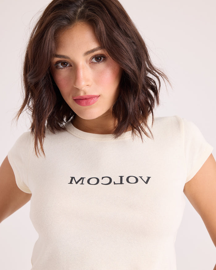A woman with shoulder-length brown hair is wearing the Volcom Women's Flip Side 1991 Mid Tee in vintage white, a fitted cropped tee featuring the word "VOLCOM" printed backward. She faces the camera directly against a plain background.
