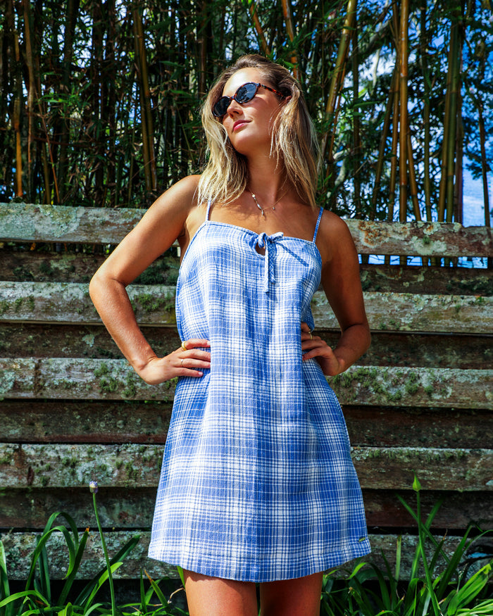 A woman poses outdoors on a sunny day wearing Volcom's Women's Folkin Stone Dress—an A-line mini with a blue plaid pattern—and sunglasses, standing in front of a wooden fence and tall bamboo.