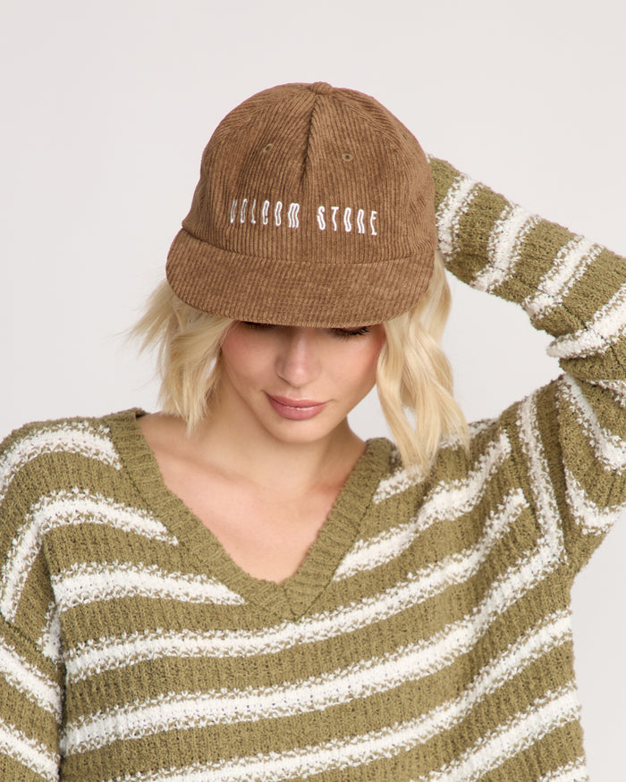 A person with short blonde hair wears a brown Volcom Women's Full Stone Dad Hat with "VOLCOM STONE" on the front and an olive-green striped cotton sweater. Their head is slightly down, one hand touching the cap's side.