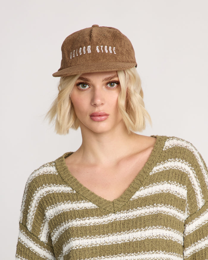 A woman with short blonde hair wears the Volcom Women's Full Stone Dad Hat in brown corduroy with white embroidery and an olive-green and white striped V-neck sweater, looking at the camera against a plain background.