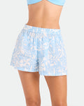 A person is shown from waist to mid-thigh wearing Volcom Women's Funtastical Gauze Shorts—light blue relaxed fit with a white floral pattern and an elasticated waist—paired with a matching light blue cropped top against a plain background.