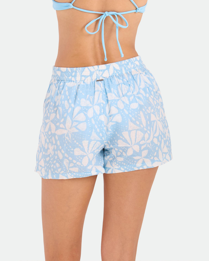 A woman is shown from the back wearing Volcom's Women's Funtastical Gauze Shorts in light blue with a floral pattern, relaxed fit, and elasticated waist, paired with a matching bikini top against a plain white background.