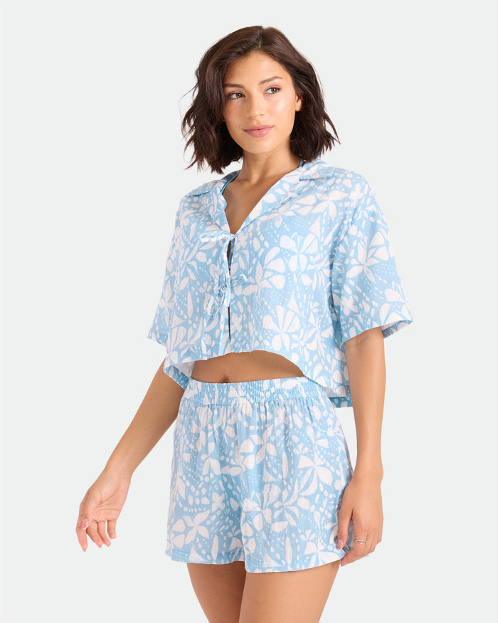 A woman wears Volcom's Women's Funtastical Gauze Shorts—high-waisted, elasticated, double-gauze cotton with a white floral print—paired with a matching light blue crop top, standing against a plain light background.