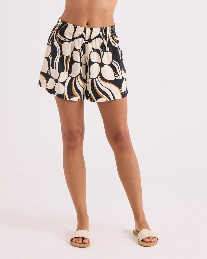 A model showcases the Volcom Women's Funtastical Shorts—high-waisted pull-on shorts featuring an elastic waist and a relaxed fit with an abstract black, white, orange, and beige print. Paired with beige sandals, the look is styled against a plain white background for a comfortable yet stylish ensemble.