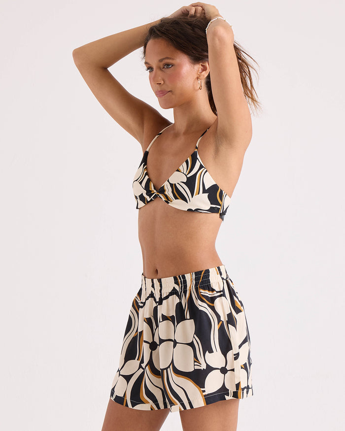 A woman poses with her arms raised against a plain light background, wearing the Volcom Women's Funtastical Shorts—a relaxed fit, printed pull-on style in black, white, and yellow with an elastic waistband for comfort and style—paired with a matching printed bralette.