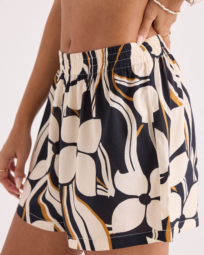 Wearing the Volcom Women's Funtastical Shorts, which feature a relaxed fit and high-waisted design with a bold, abstract floral print in cream, black, and brown tones, a person poses with one hand on their hip while accessorizing with a beaded bracelet.