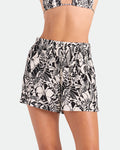A woman wears Volcom Women's Funtastical Viscose Shorts in a black ecru tropical leaf print with an elastic waistband, paired with a matching bikini top against a plain light-colored background.