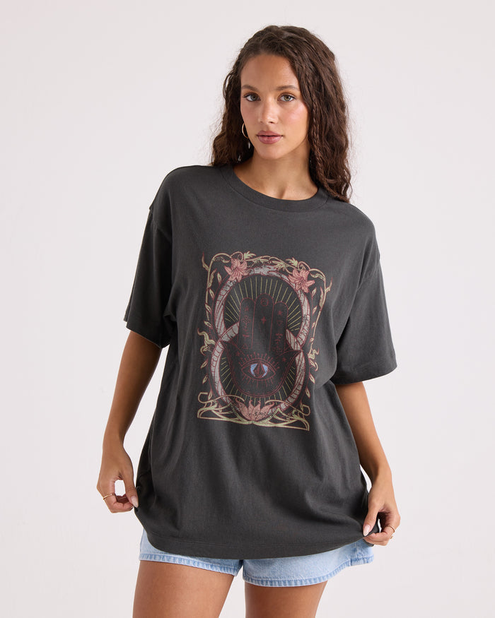 A woman with long curly hair wears Volcom’s Women’s Gatekeeper Stones Throw Tee—an oversized, 100% cotton vintage black t-shirt featuring an abstract mystical design—paired with light denim shorts as she poses against a plain white background.