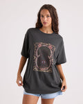 A woman with long curly hair wears Volcom’s Women’s Gatekeeper Stones Throw Tee—an oversized, 100% cotton vintage black t-shirt featuring an abstract mystical design—paired with light denim shorts as she poses against a plain white background.