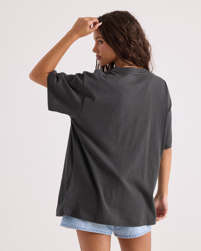 A woman with wavy brown hair stands with her back to the camera, touching her hair, wearing the Volcom Women's Gatekeeper Stones Throw Tee—an oversized vintage black t-shirt made from 100% cotton—paired with light denim shorts. The background is plain and light-colored.