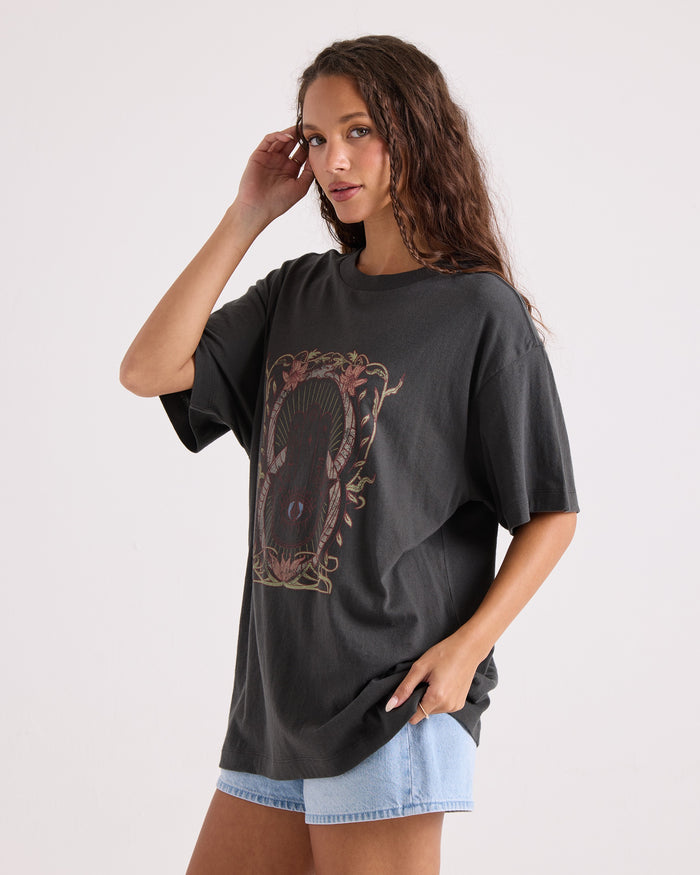 A woman with long wavy hair poses against a plain background, wearing the Volcom Women's Gatekeeper Stones Throw Tee—an oversized black vintage-style t-shirt made from 100% cotton—and light denim shorts, with one hand near her face and the other in her pocket.