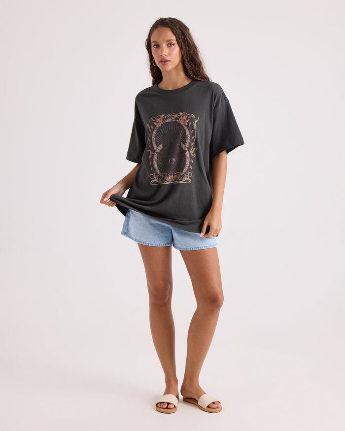 A woman stands confidently against a plain background wearing the Volcom Women's Gatekeeper Stones Throw Tee—an oversized vintage black t-shirt crafted from 100% cotton and featuring a bold, ornate graphic design. She pairs it with light denim shorts and white slide sandals, holding the side of her shirt as she gazes at the camera.