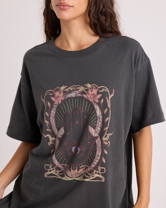 A woman wears the Volcom Women's Gatekeeper Stones Throw Tee—an oversized vintage-style black T-shirt made from 100% cotton, showcasing a mystical hand-with-eye motif at the center and surrounded by ornate floral and geometric patterns.