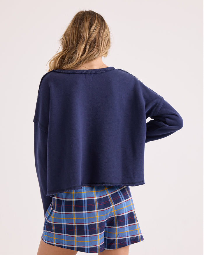A person with long hair stands facing away, wearing a relaxed fit navy blue Women's Gnarly Sweatshirt by Volcom and blue plaid shorts against a plain light background.