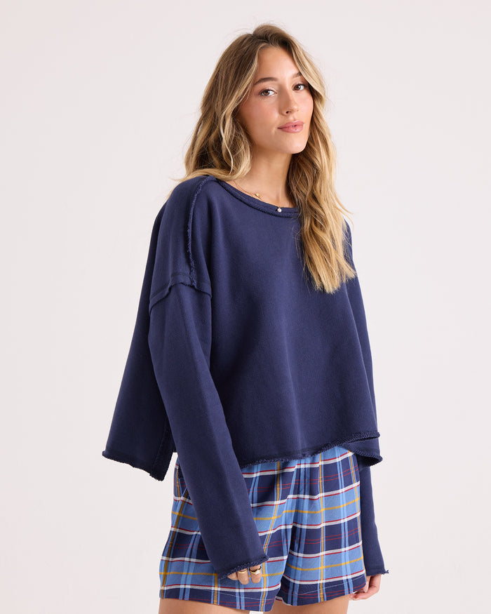 A woman with long, wavy hair wears the Volcom Women's Gnarly Sweatshirt in a relaxed fit, paired with blue plaid shorts, standing against a plain light background.