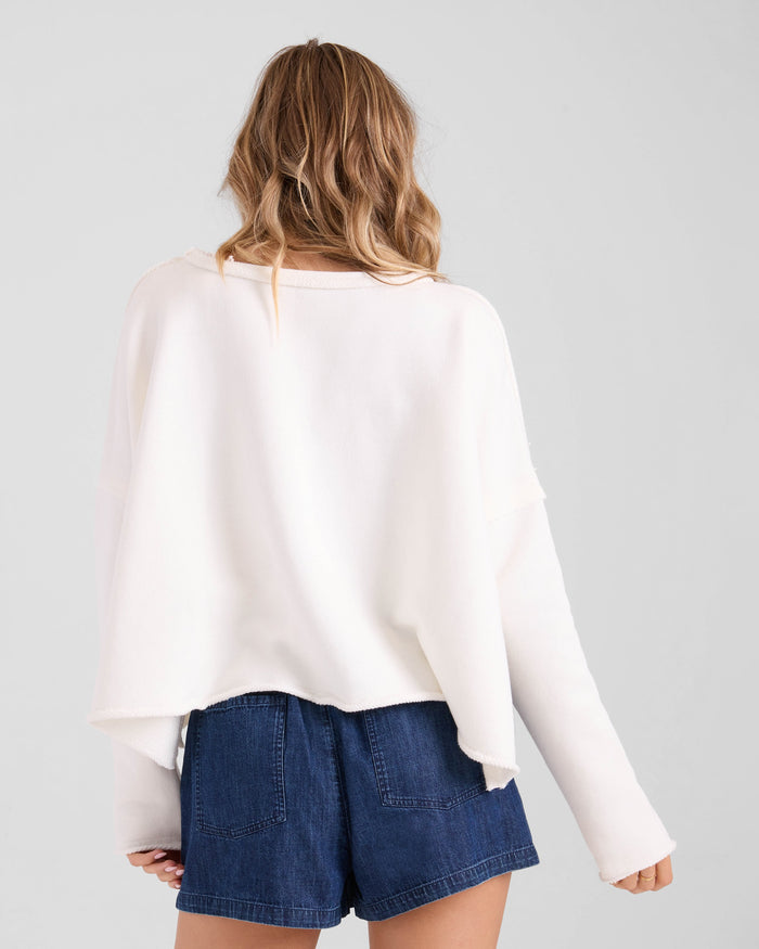 A person with wavy, light brown hair is seen from the back, wearing the Volcom Women's Gnarly relaxed fit sweatshirt paired with high-waisted dark denim shorts against a plain light background.