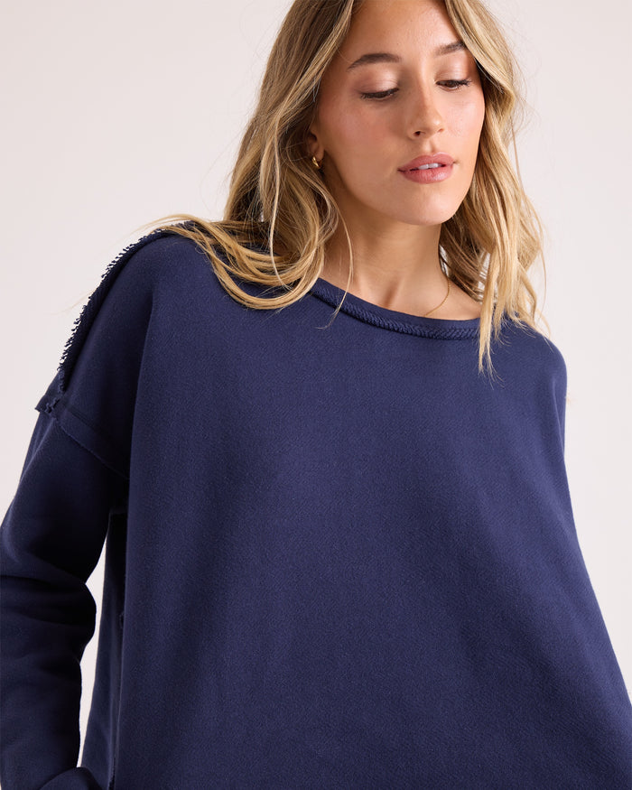 A woman with long, wavy blonde hair wears the Volcom Women's Gnarly Sweatshirt in a relaxed fit navy blue dolman style, looking downward with a neutral expression against a plain light background.