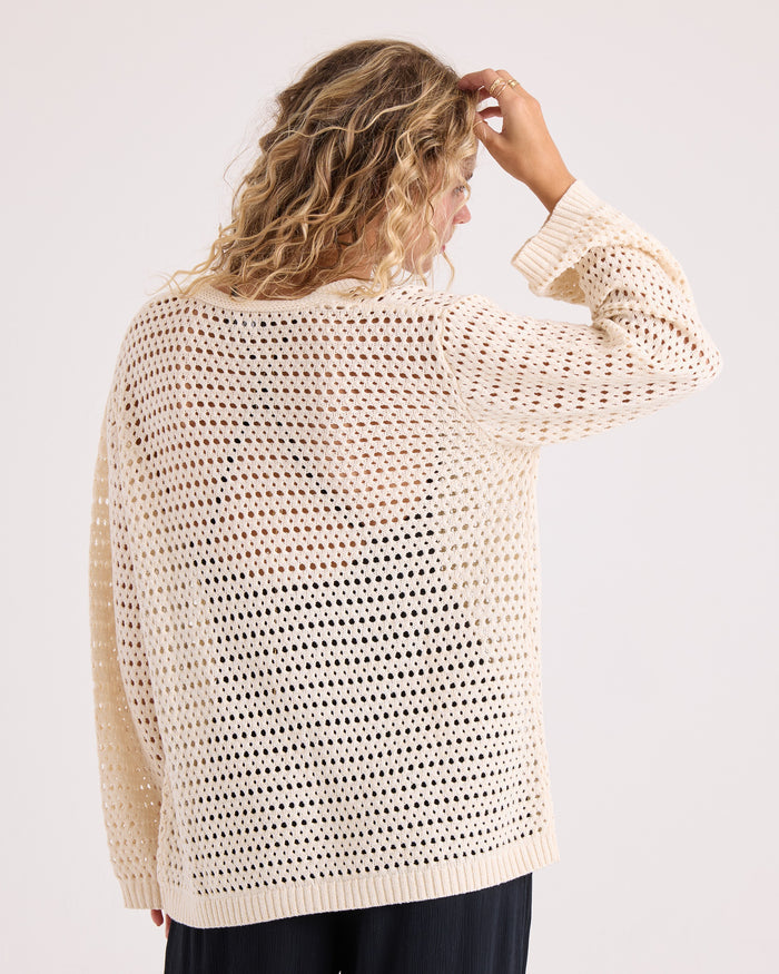 A person with wavy blonde hair stands with their back to the camera and hand raised to their head against a plain background, wearing the Volcom Women's Go Go Wrap—a loose, cream-colored, open-knit sweater top crafted from a soft cotton-acrylic blend—paired with black pants.