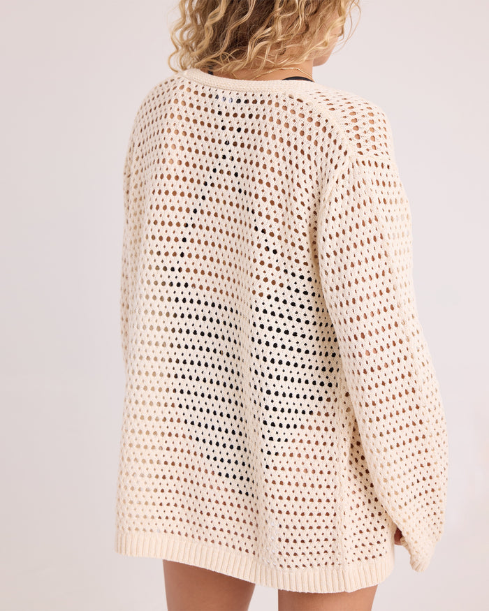 A person with curly blonde hair is shown from the back, standing against a plain, light background and wearing the Volcom Women's Go Go Wrap—a loose, cream-colored, open-knit sweater top crafted from a soft cotton-acrylic blend.