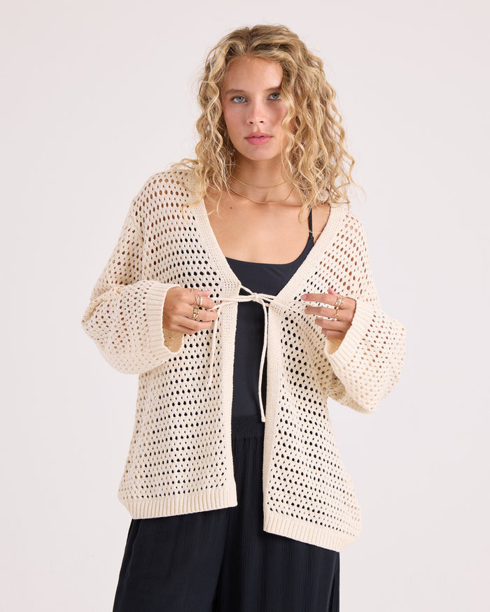 A woman with curly blonde hair wears the Volcom Women's Go Go Wrap, a cream-colored, open-knit tie-front cardigan, over a black top and pants, standing against a plain background.