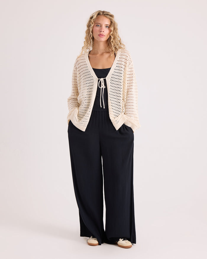 Wearing the Volcom Women's Go Go Wrap, a person with long curly hair stands against a plain light background with their hands in their pockets, layering the cream cotton-acrylic blend cardigan over a black top and loose black pants.