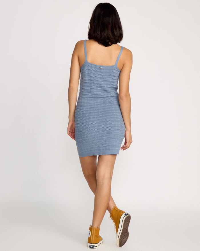 A woman with short dark hair wears the Volcom Women's Gurrl Gang Dress in Carolina Blue, a ribbed, sleeveless knit style with a body-skimming fit, paired with yellow high-top sneakers as she walks away from the camera against a white background.