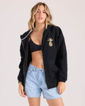 A woman wearing the Volcom Women's Haleiwa black hoodie with a gold pineapple graphic, a black sports bra, and light blue denim shorts stands against a plain light background.