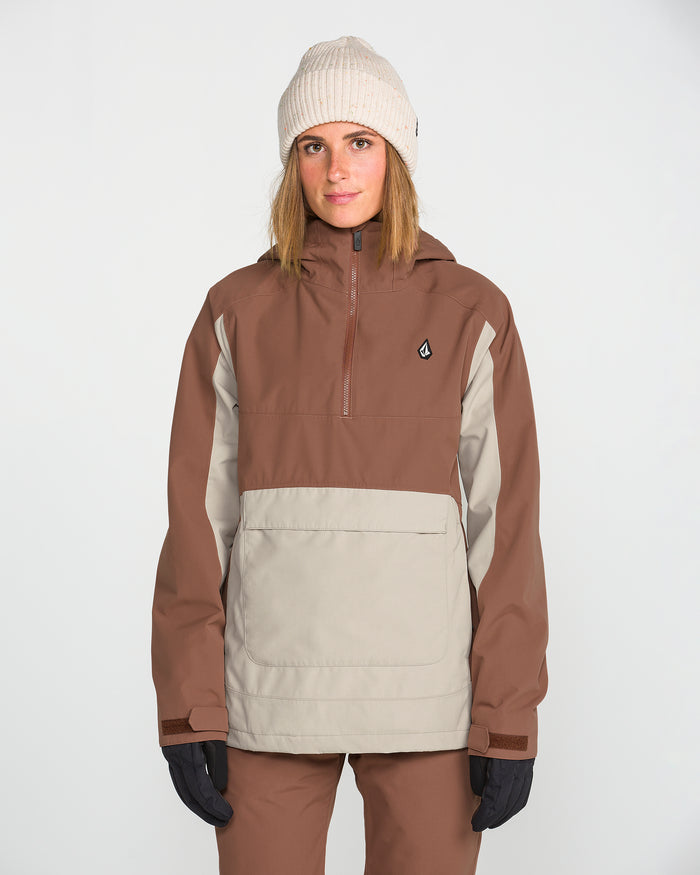 A person wearing a beige beanie, brown pants, black gloves, and the Volcom Women’s Harlan Pullover—a brown and beige jacket with a recycled shell—stands facing forward against a plain white background.