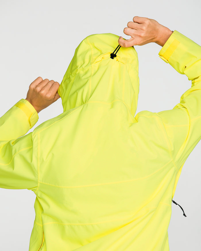 A person seen from behind adjusts the hood drawstring of the Volcom Women’s Harlan Pullover waterproof jacket with one hand, while holding the side of the hood with the other.