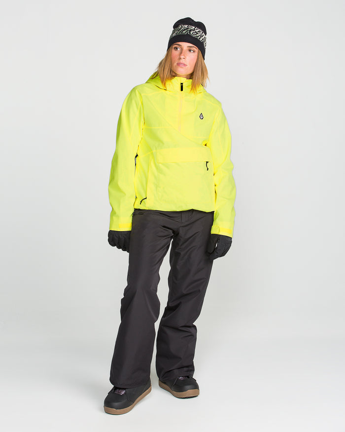 A person wearing the Volcom Women’s Harlan Pullover—a bright yellow waterproof jacket made from recycled shell material—paired with black snow pants, black gloves, black boots, and a patterned black beanie stands against a plain light background.