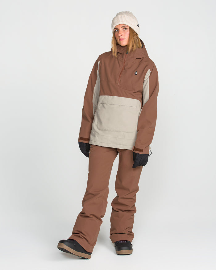 A person stands against a plain white background wearing the Volcom Women’s Harlan Pullover—a tan and brown waterproof jacket with a recycled shell—paired with matching pants, black gloves, a beige knit beanie, and black winter boots.