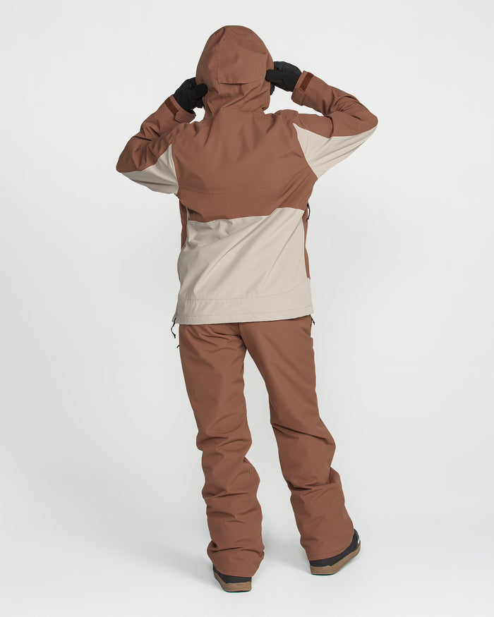 A person stands on a white background with both hands raised to adjust the hood of their Volcom Women’s Harlan Pullover, a brown and beige waterproof jacket. They are facing away from the camera.