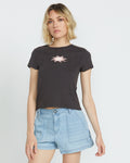 A woman with wavy blonde hair wears the Volcom Women's Have A Clue Tee, a black cotton rib shirt with a pink chest graphic, and light blue denim shorts, standing against a plain white background.
