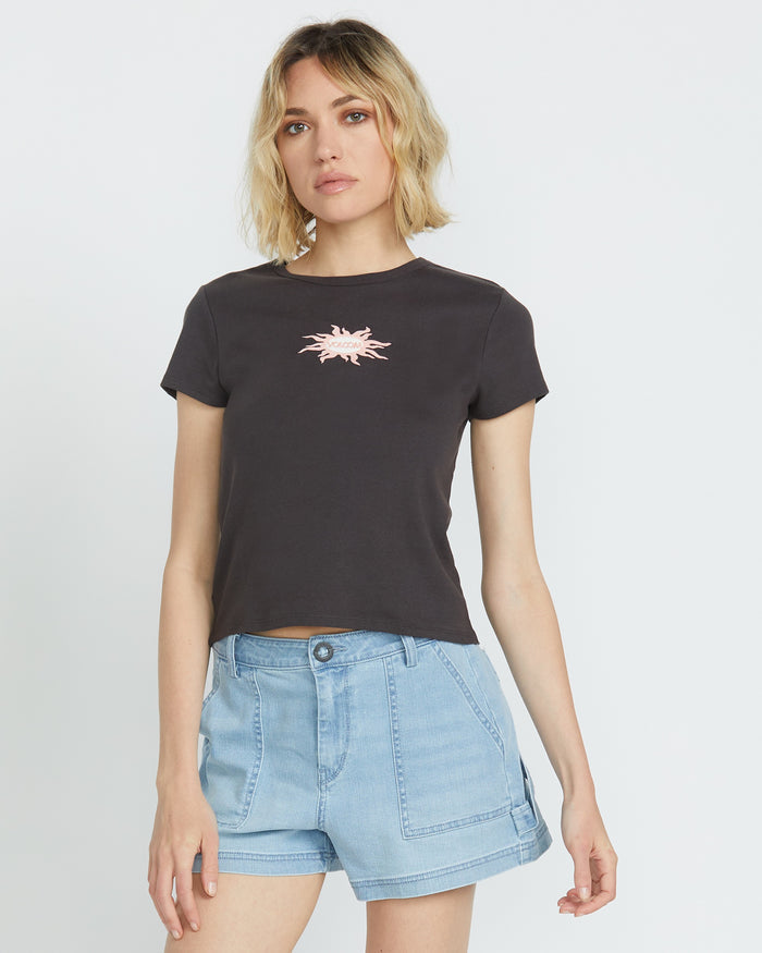 A woman with wavy blonde hair wears the Volcom Women's Have A Clue Tee, a black cotton rib shirt with a pink chest graphic, and light blue denim shorts, standing against a plain white background.