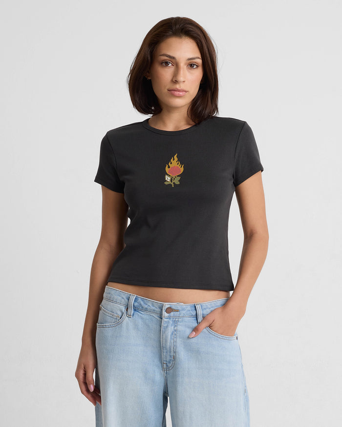 A woman with straight brown hair wears the Volcom Women's Have A Clue Tee, a fitted black t-shirt with a small flaming torch graphic on the chest, paired with light blue jeans. She stands against a plain white background with a neutral expression.